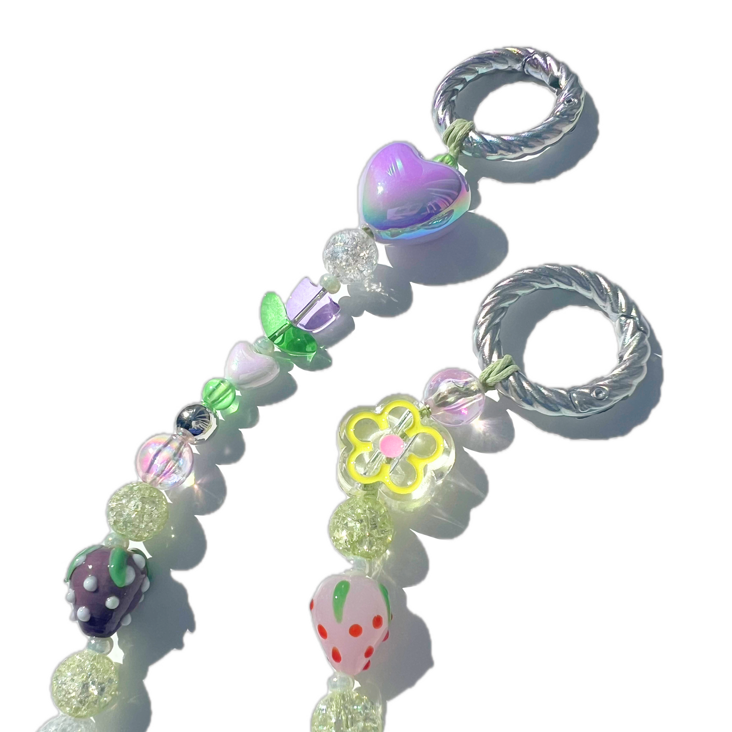 Close-up of a handmade beaded bag charm featuring pastel purple, green, and yellow accents with heart, flower, and strawberry beads. The iridescent charm sparkles in the sunlight, attached to twisted silver clasps.