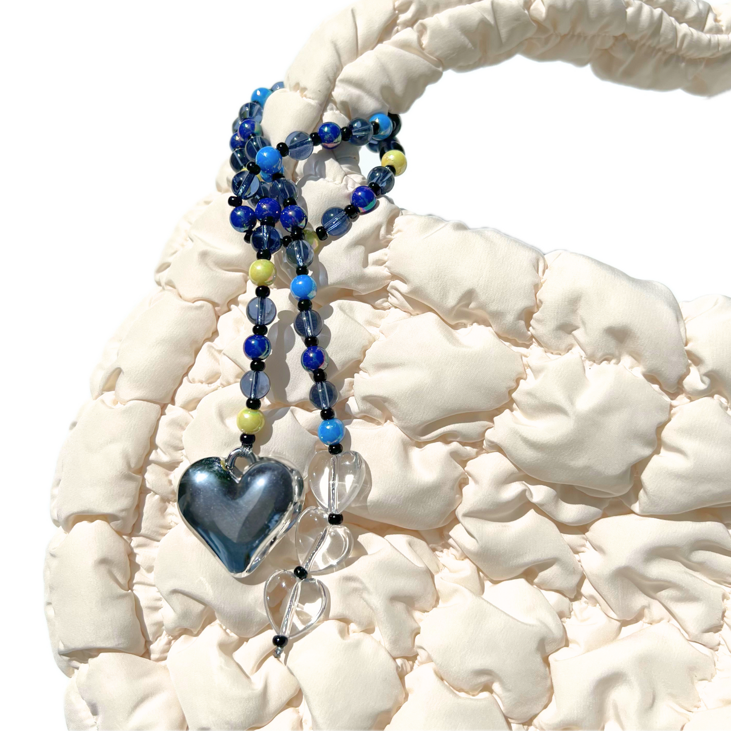 The Chrome Silver Heart Bag Charm with midnight blue and clear crystal beads clipped to a textured cream handbag, featuring a reflective chrome heart pendant and translucent clear heart accents.