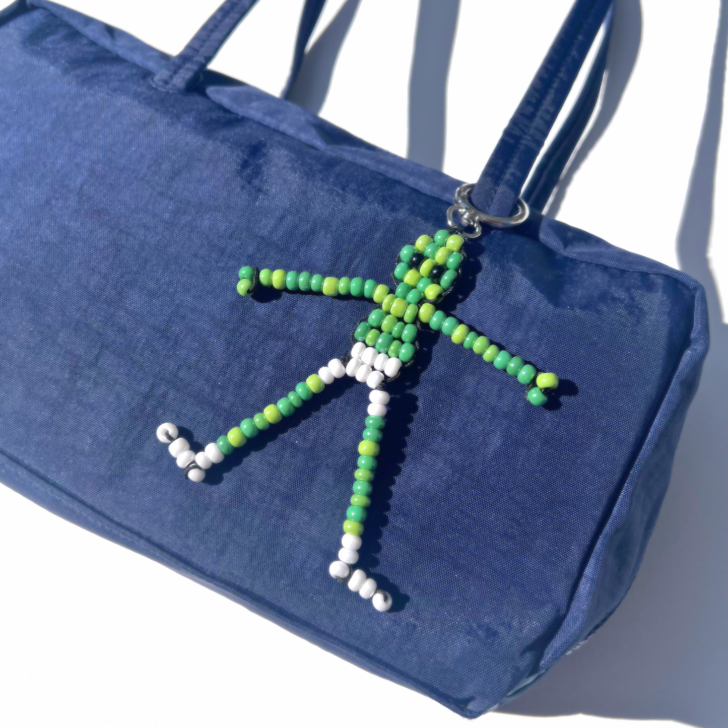 The Beaded Green Martian Bag Charm clipped to a navy blue handbag, showcasing the playful alien figure made of green, white, and black beads with silver hardware.