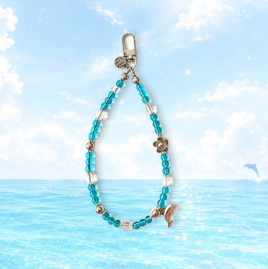 Aqua dolphin beaded phone wristlet charm with a silver dolphin pendant, perfect vacation accessory styled against a sunny blue ocean and sky