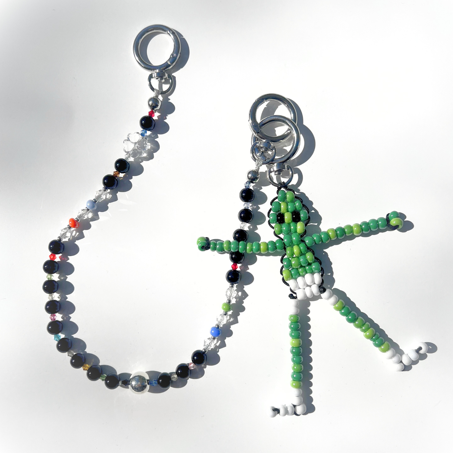 The Black Orbit Beaded Bag Charm paired with a green beaded alien keychain, both lying on a white surface with sunlight reflecting off the beads.