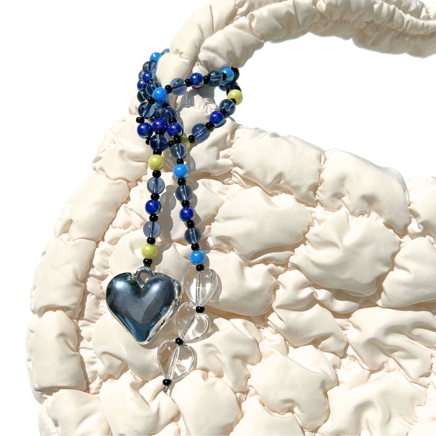 The Chrome Silver Heart Bag Charm with midnight blue and clear crystal beads clipped to a textured cream handbag, featuring a reflective chrome heart pendant and translucent clear heart accents.