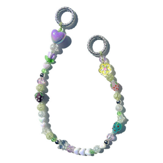 Full view of a pastel garden-themed beaded bag charm laid flat, showing whimsical details like hearts, strawberries, glass beads, and a yellow flower. Attached to two twisted silver loops.