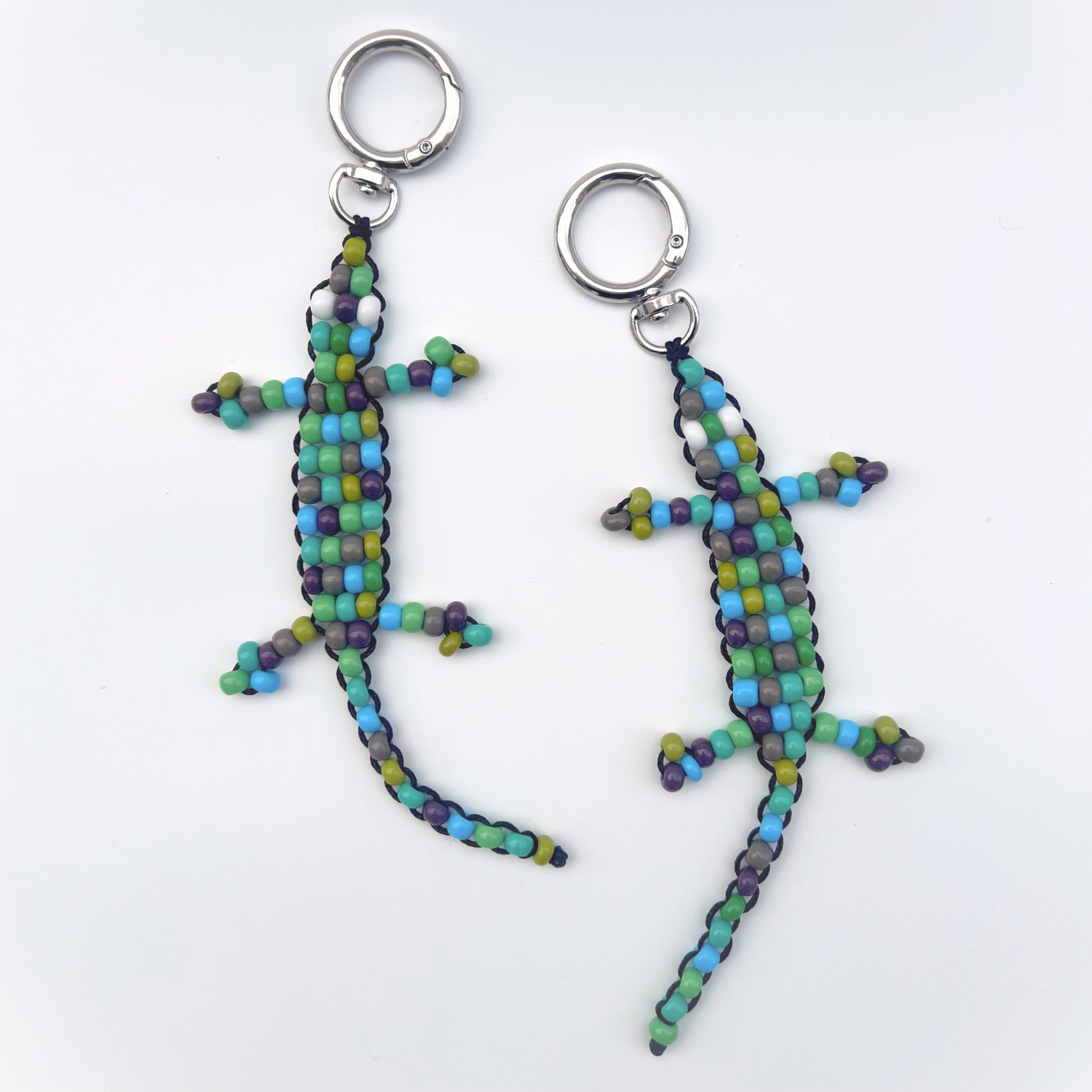 Two handmade green, blue, and grey beaded gecko bag charms laid flat on a white background with metal keyrings.