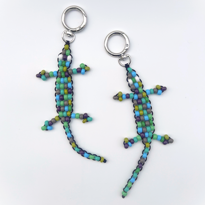 Two handmade green, blue, and grey beaded gecko bag charms laid flat on a white background with metal keyrings.