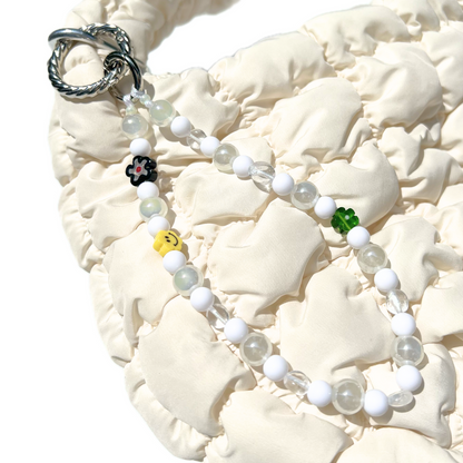 Luminous white eclectic beaded charm clipped to a cream quilted bag, featuring milky glass and clear beads with playful accents including a yellow smiley, green and black flowers. Handmade with glass, Czech, millefiori, and acrylic beads.