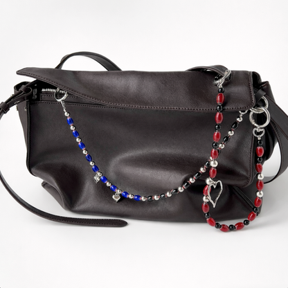 Dark brown leather shoulder bag photographed on a white background, styled with two beaded bag charms—one in blue, black, and silver draped across the front, and one in red, black, and silver with a heart-shaped charm hanging on the right side.