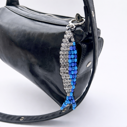 Blue and silver beaded sardine fish bag charm attached to a black leather handbag, shown hanging from a zipper pull.