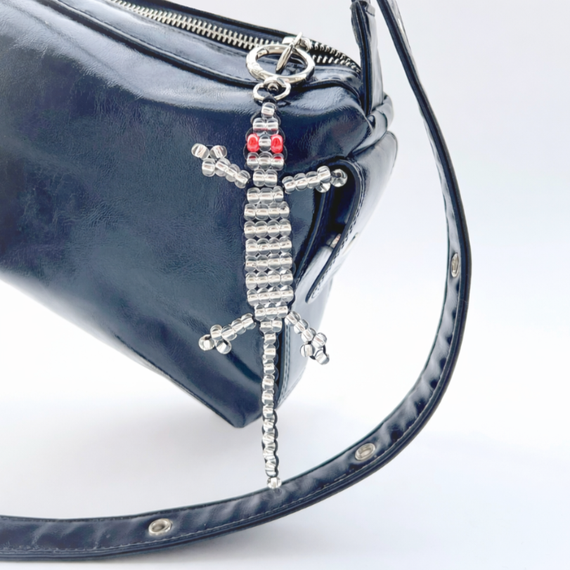 Silver beaded gecko bag charm with red eyes attached to a black leather handbag, shown hanging from a zipper ring.
