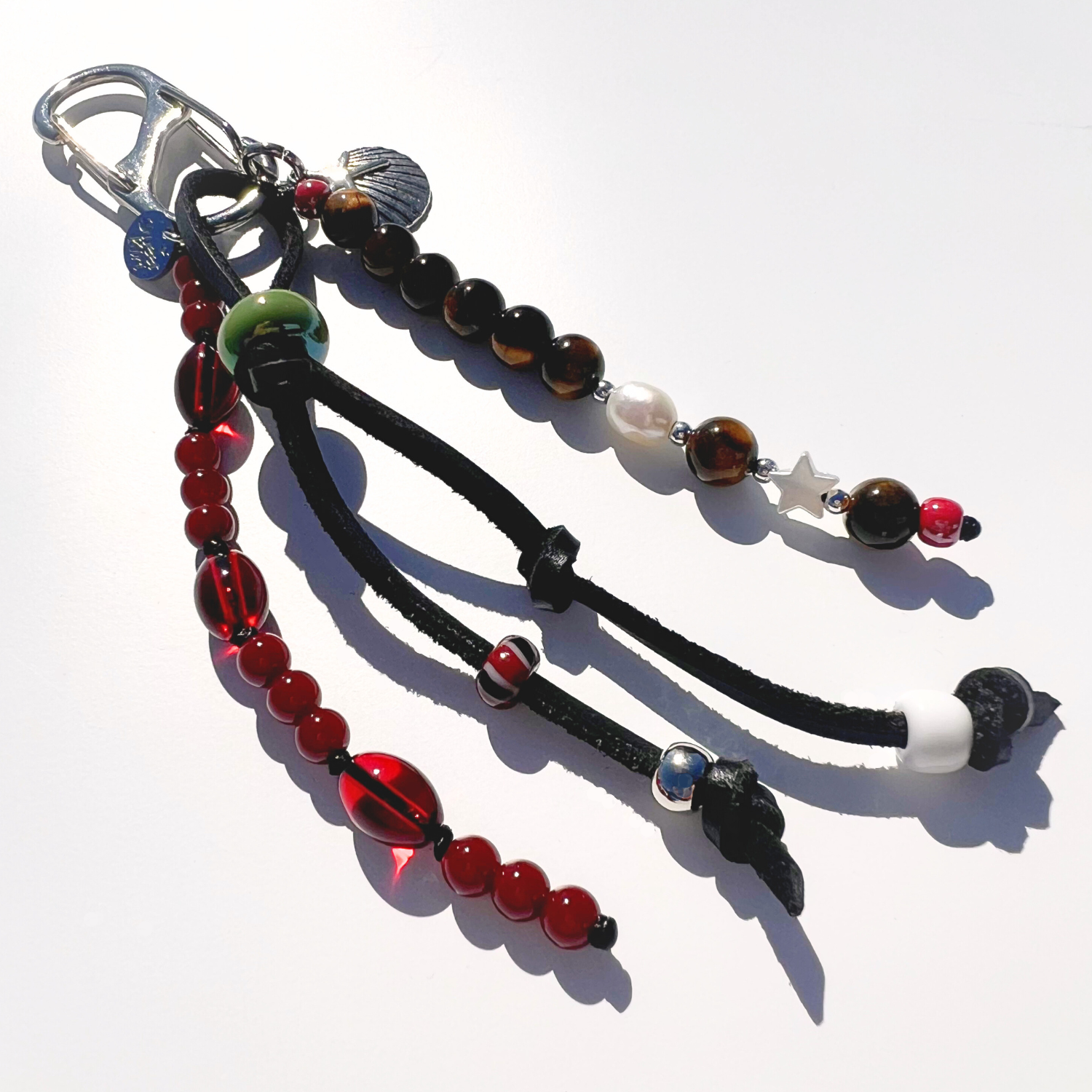 Close up of Mariner Relic Tiger Eye & Crimson bag charm showing tiger eye stones, red glass beads, leather cord knots, and starfish shell pendent 