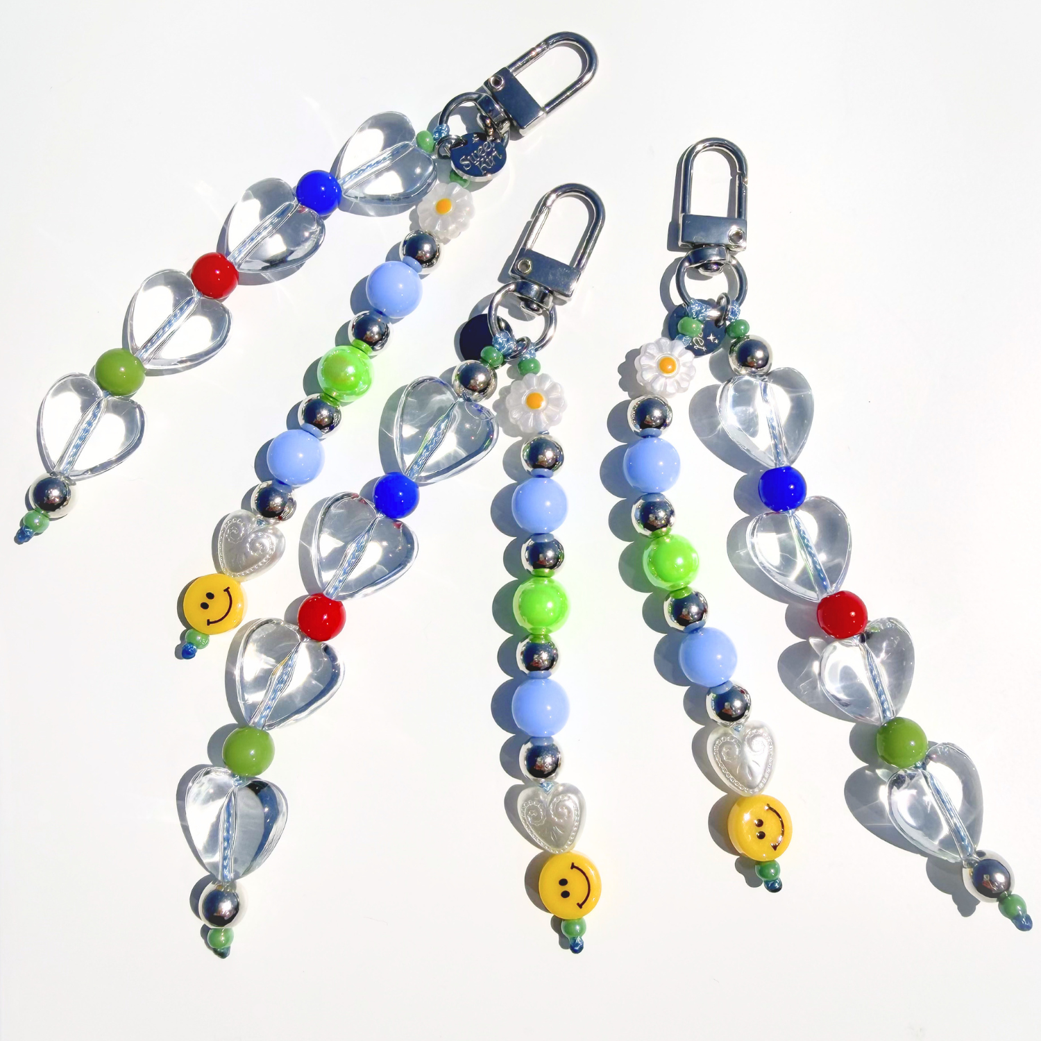 Close-up detail shot of handmade beaded bag charms from SweetKiri. Featuring clear heart beads, yellow smiley, daisy flower, and silver keyring clasp