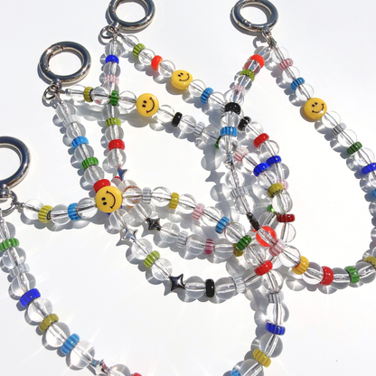 A close-up detail shot of multiple Solar Joy Beaded Bag Charm showing clear beads, multicolor glass rings, silver stars, and a yellow smiley bead catching the light on a white surface.