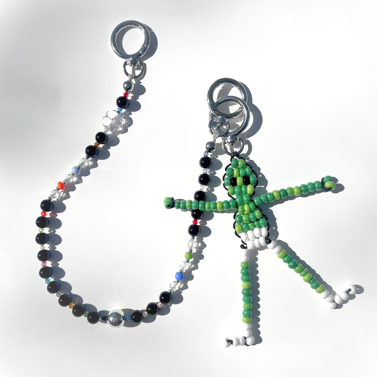 The Black Orbit Beaded Bag Charm paired with a green beaded alien keychain, both lying on a white surface with sunlight reflecting off the beads.