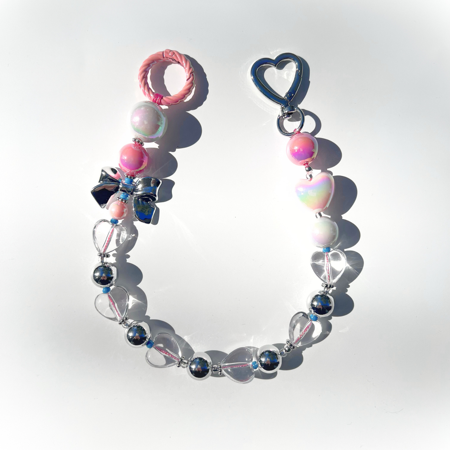 A single Kawaii Pink Beaded Bag Charm featuring pastel pink, iridescent white, and silver beads with heart and bow details, displayed on a white background under bright sunlight.