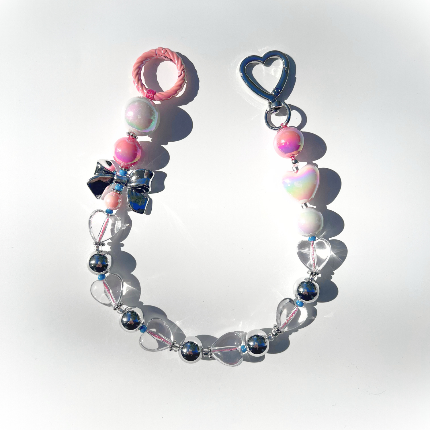 A single Kawaii Pink Beaded Bag Charm featuring pastel pink, iridescent white, and silver beads with heart and bow details, displayed on a white background under bright sunlight.